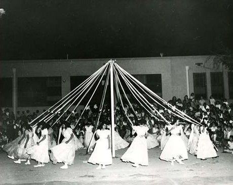 A black and white photograph of young girls in matching dresses dancing around a maypole.