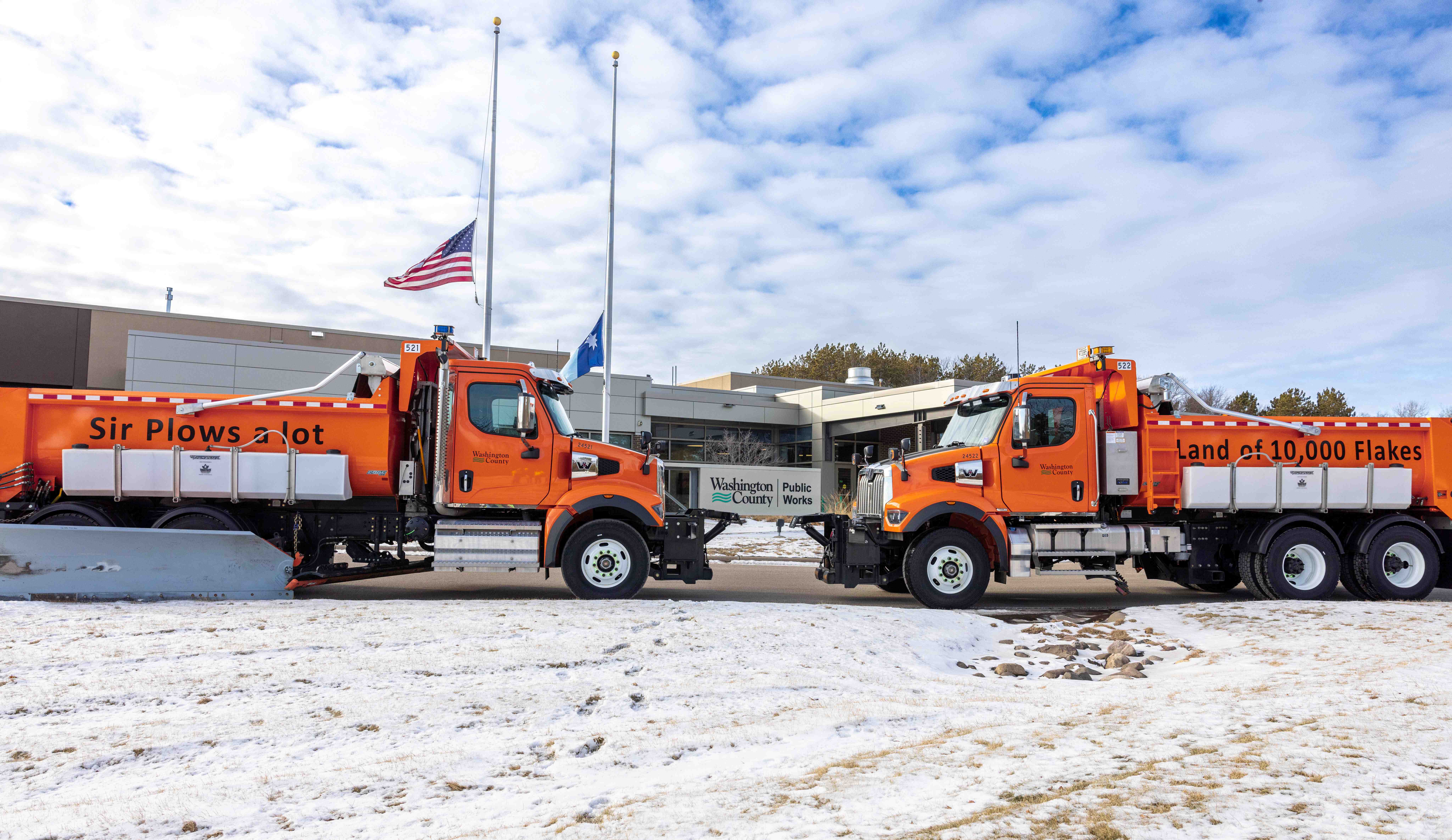 Two orange snowplow trucks parked side by side in front of a beige building under a clear blue sky.