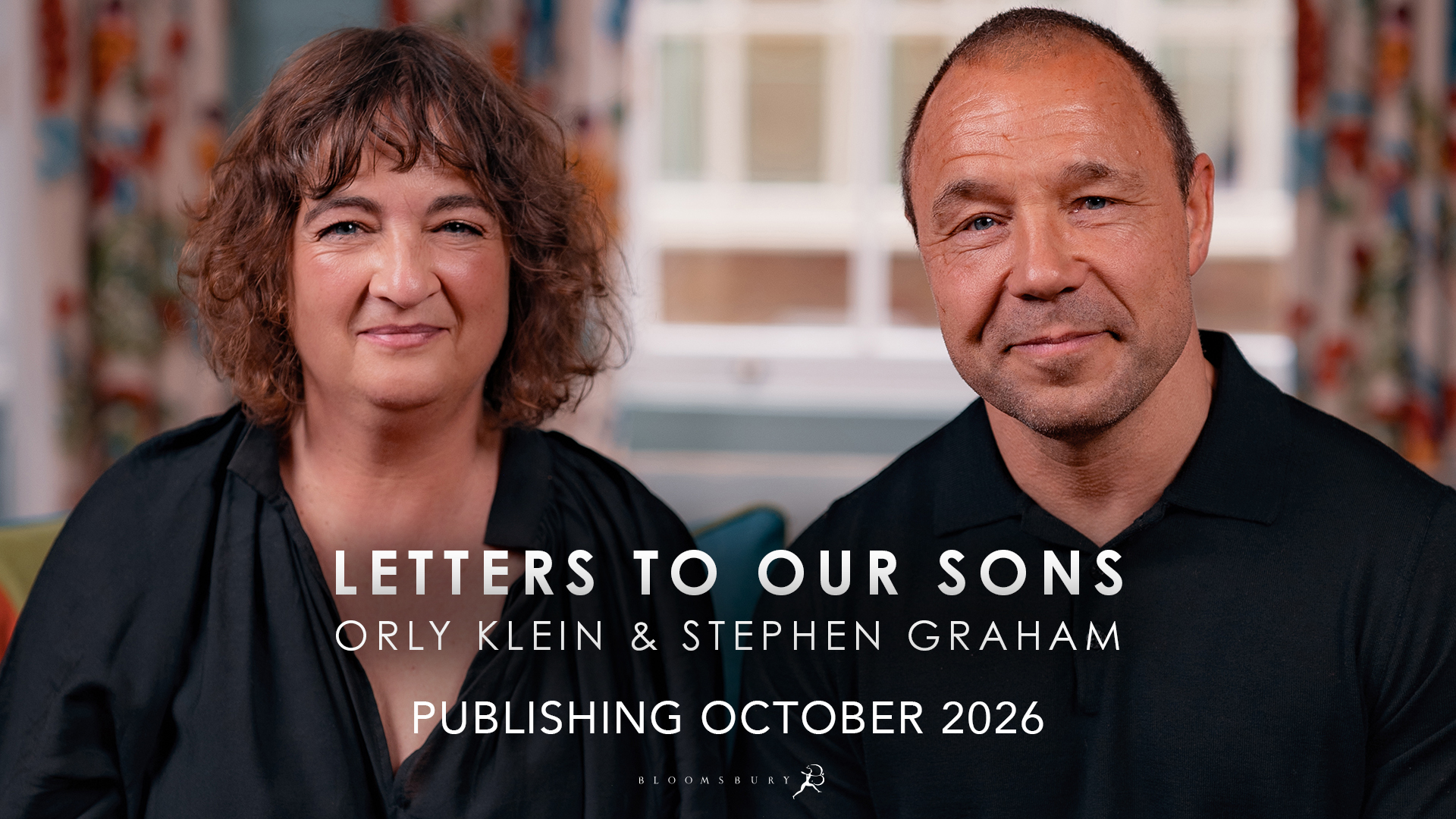 Authors Orly Klein and Stephen Graham sit side by side on a colourful couch in a room with patterned curtains and a window behind them. Orly has dark curly hair, wearing a black blouse and striped trousers. Stephen sits on the right wearing a black long sleeve shirt and dark trousers. Both are facing the camera.