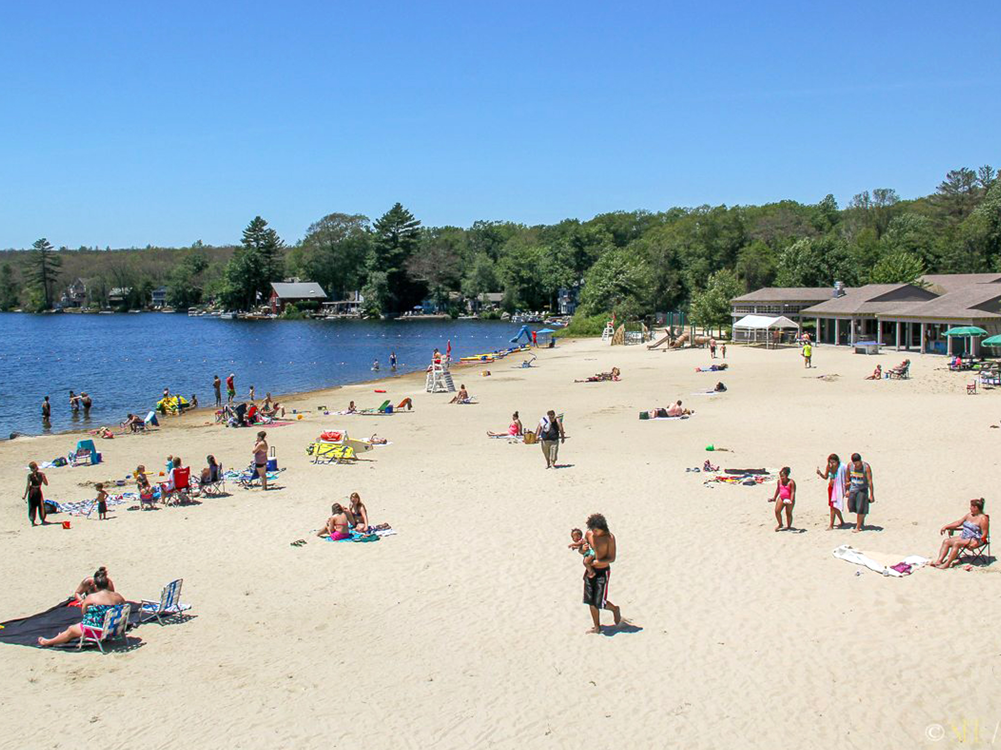 A photo of beachgoers at Spring Lake Beach in Burrillville, RI. People are laying on towels on the sand, walking on the beach, and swimming in the lake. It is a sunny day with blue skies.
